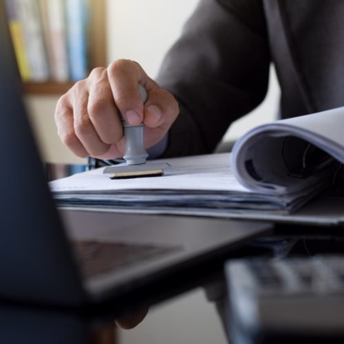 Businessman in suit hand stamping rubber stamp on document in file folder with laptop computer on the desk at office. Authorized allowance permission approval concept.