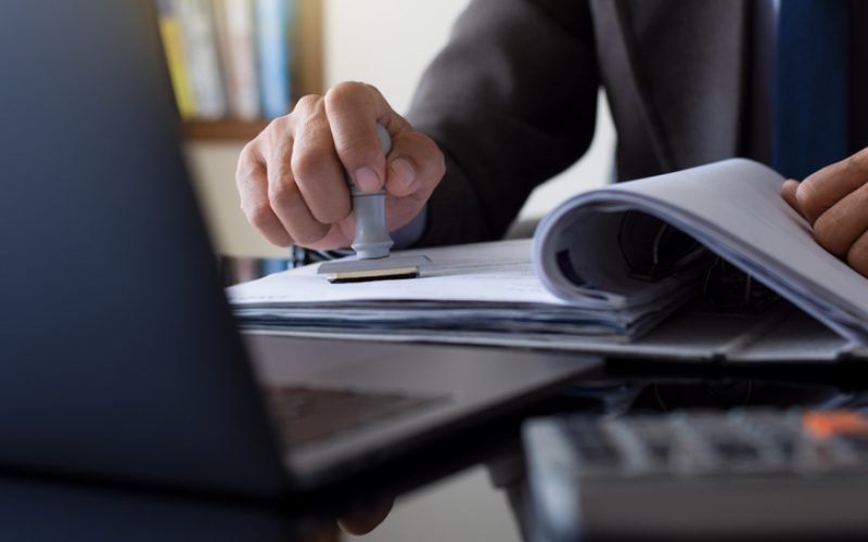 Businessman in suit hand stamping rubber stamp on document in file folder with laptop computer on the desk at office. Authorized allowance permission approval concept.