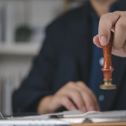 Businessman hand holding stamp seal, approval legal document. Official authorization, contract agreement verification. Lawyer notary work office desk, finalize paperwork, confidential file, laptop.