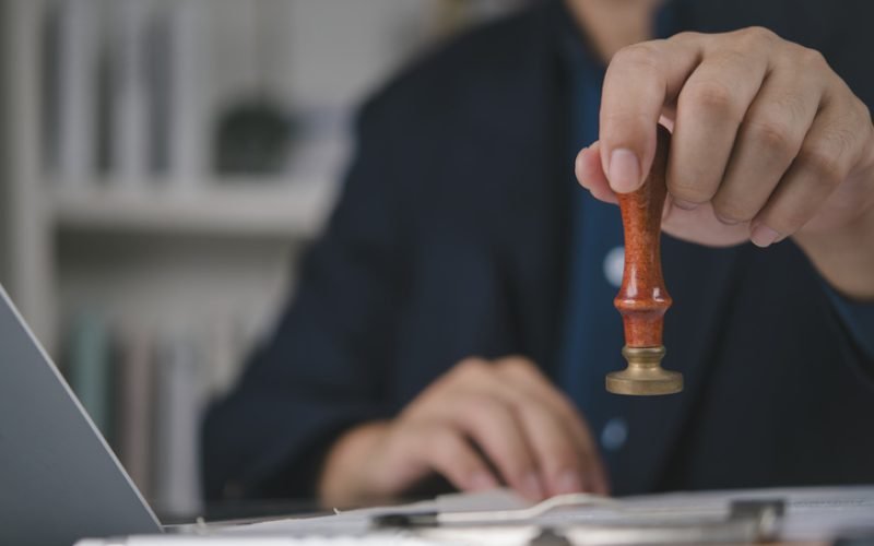 Businessman hand holding stamp seal, approval legal document. Official authorization, contract agreement verification. Lawyer notary work office desk, finalize paperwork, confidential file, laptop.