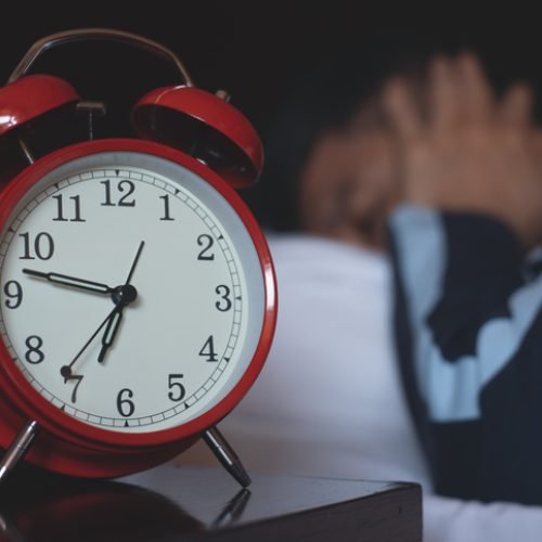 Drowsy man sleeping with red alarm clock on bed side table in his bedroom, waking up in early morning, close up
