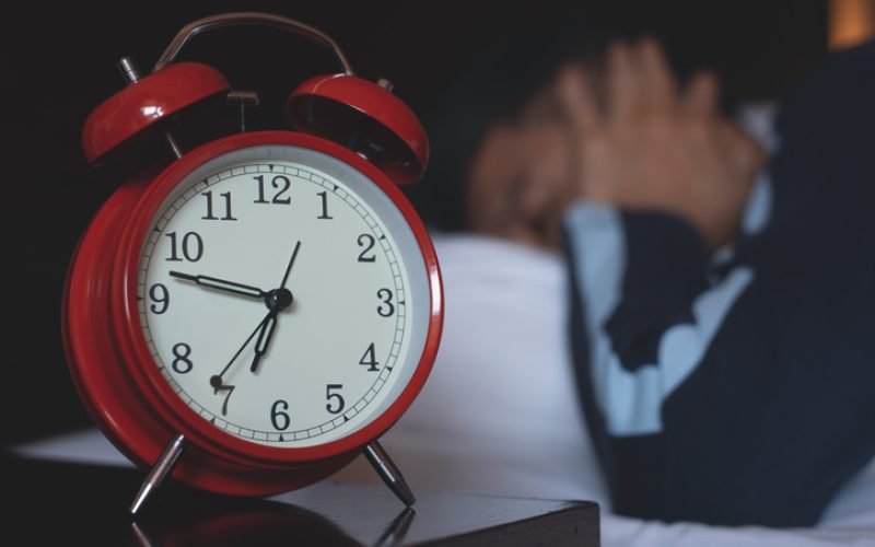 Drowsy man sleeping with red alarm clock on bed side table in his bedroom, waking up in early morning, close up
