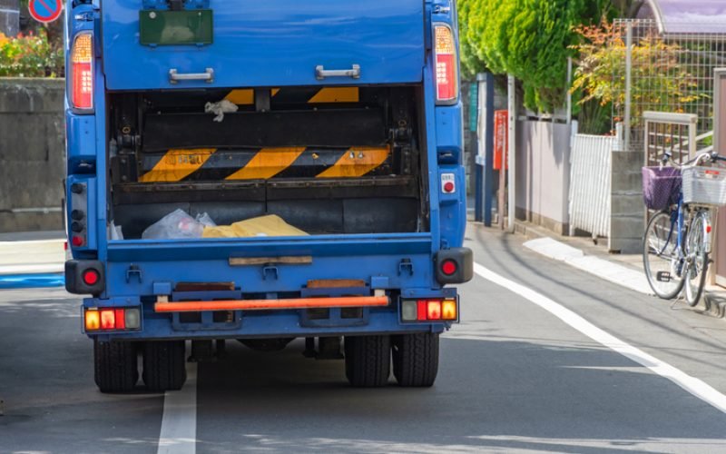 The photo captures the rear view of a garbage truck as it operates within a city environment. This might include details of the truck's mechanisms, collected waste, or the urban surroundings.