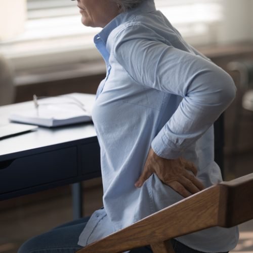 Cropped unhealthy middle aged senior woman employee massaging stiff back muscles, suffering from sudden pain, sitting at table doing computer work, feeling strong lumbar ache at modern home office.