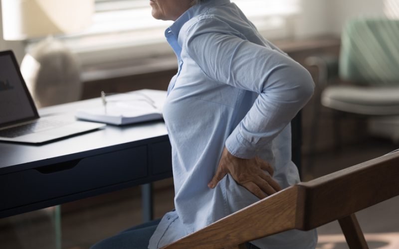 Cropped unhealthy middle aged senior woman employee massaging stiff back muscles, suffering from sudden pain, sitting at table doing computer work, feeling strong lumbar ache at modern home office.