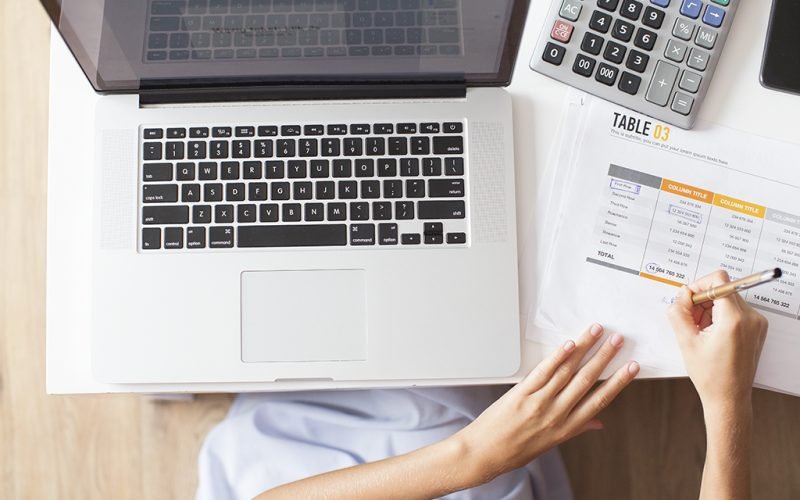 Cropped view of business woman hands making notes on document with table and working on laptop computer on desk. Top view.