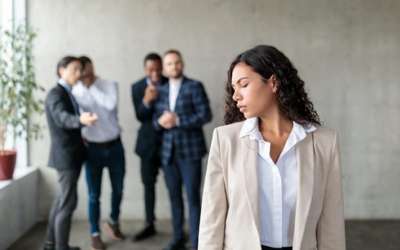 Sexism. Group Of Businessmen Whispering Behind Back Of Victimized Latin Businesswoman In Modern Office. Females Discrimination Problem, Workplace Bullying Concept. Panorama, Selective Focus