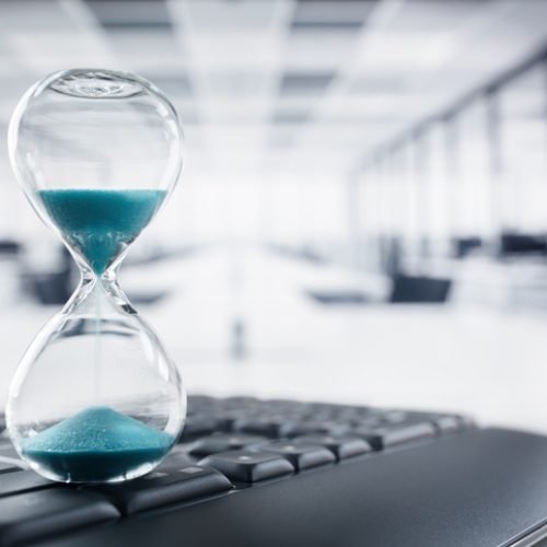 Hourglass on top of laptop computer keyboard on desk in large office concept for time management and countdown to deadline