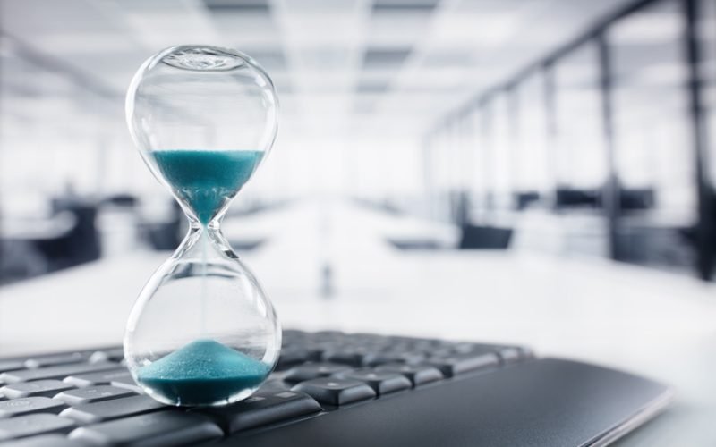 Hourglass on top of laptop computer keyboard on desk in large office concept for time management and countdown to deadline