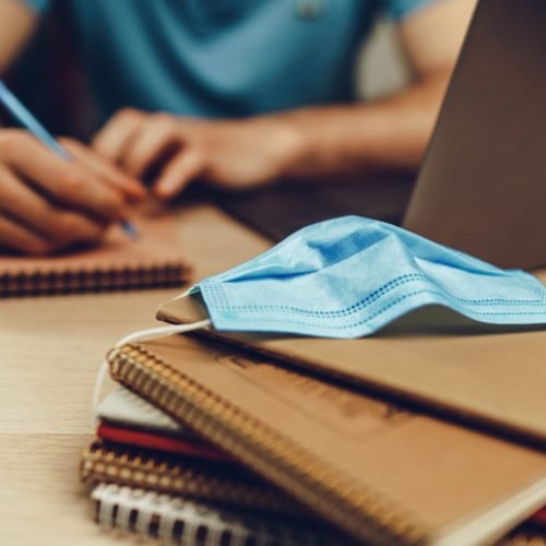 Male student studies at home during quarantine. Close up of a wooden table with laptop, notebooks and medical mask