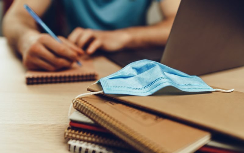 Male student studies at home during quarantine. Close up of a wooden table with laptop, notebooks and medical mask